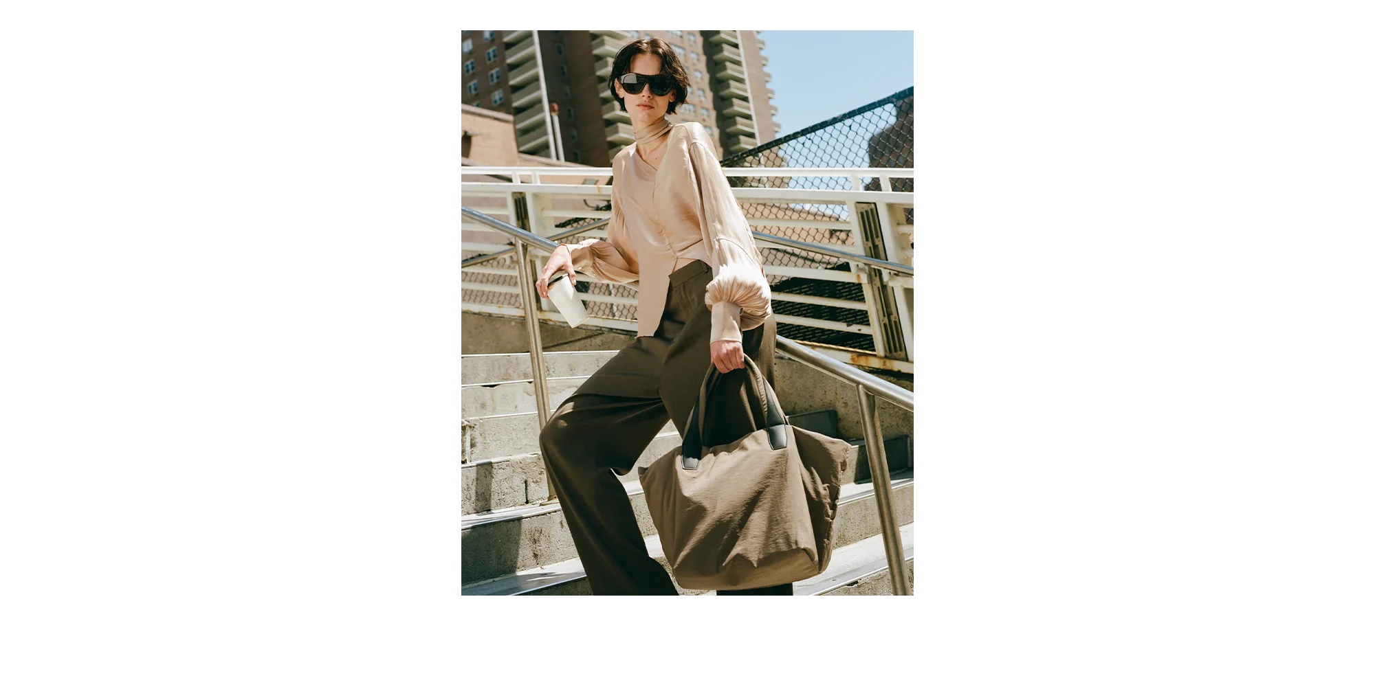 Woman standing on a metal staircase holding a mink medium tote bag, wearing sunglasses and a light-colored top.