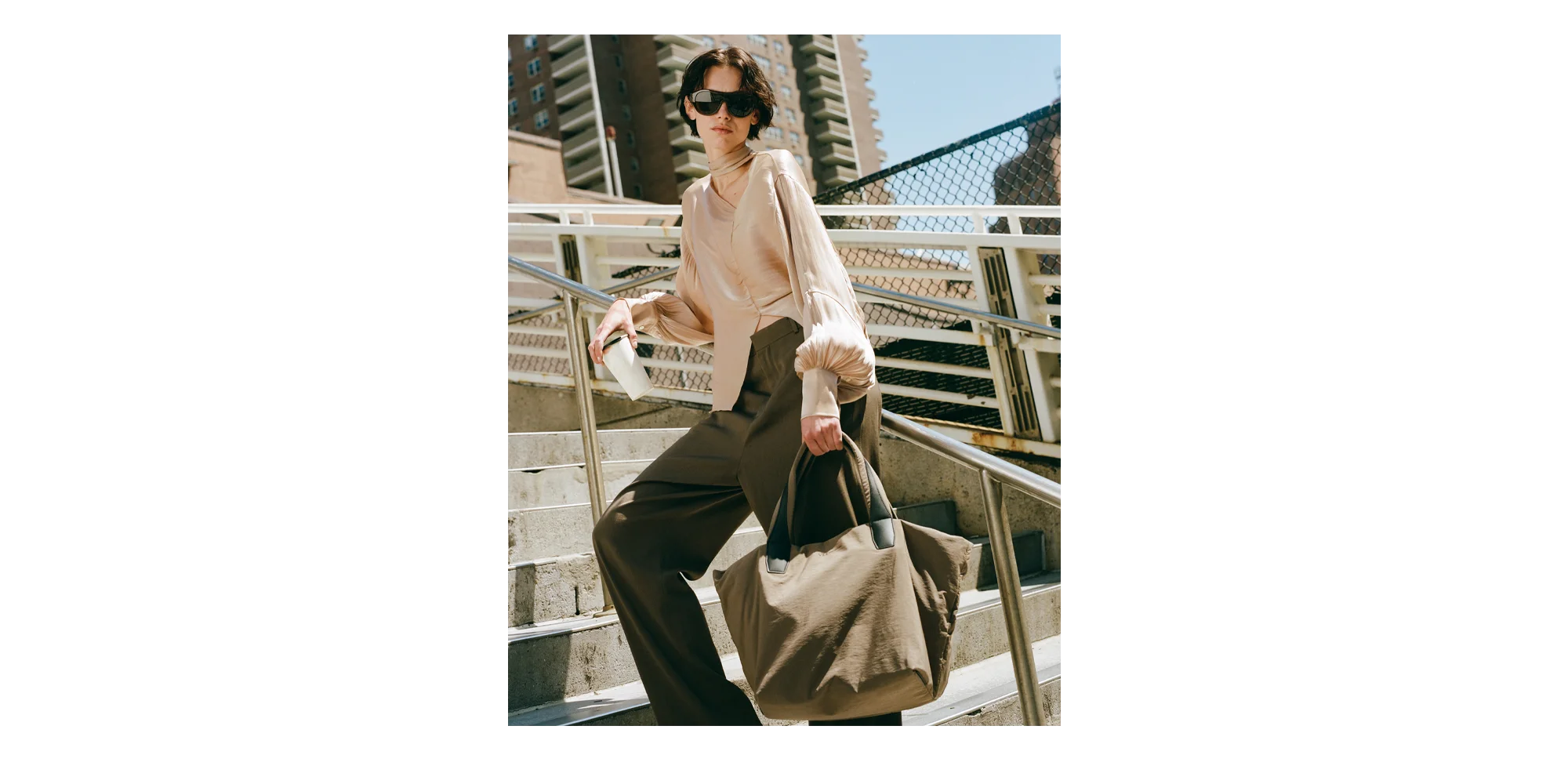 Woman standing on a metal staircase holding a mink medium tote bag, wearing sunglasses and a light-colored top.