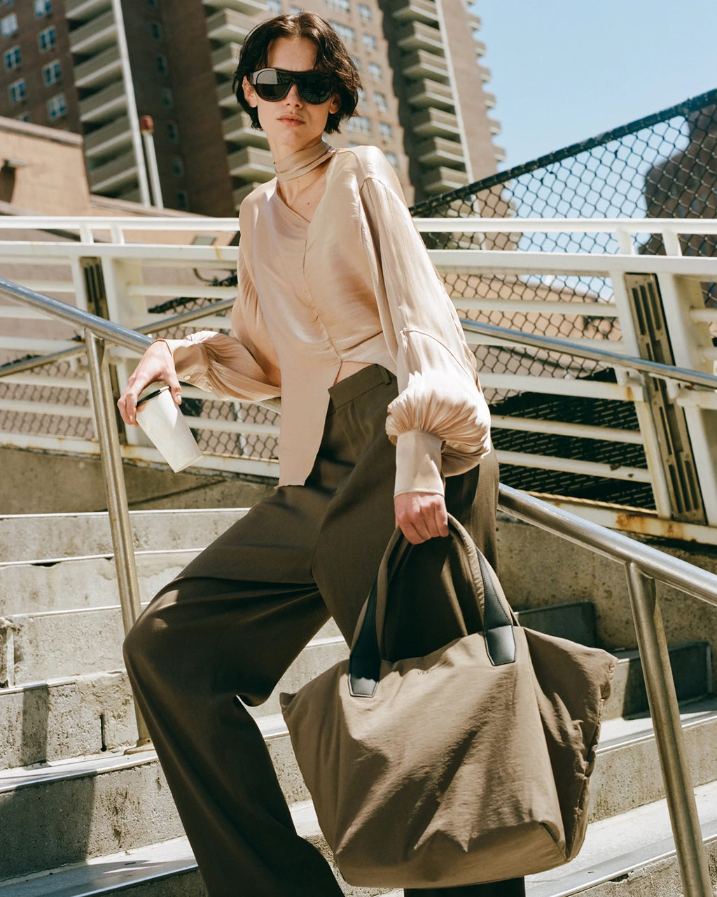 Woman standing on a metal staircase holding a mink medium tote bag, wearing sunglasses and a light-colored top.
