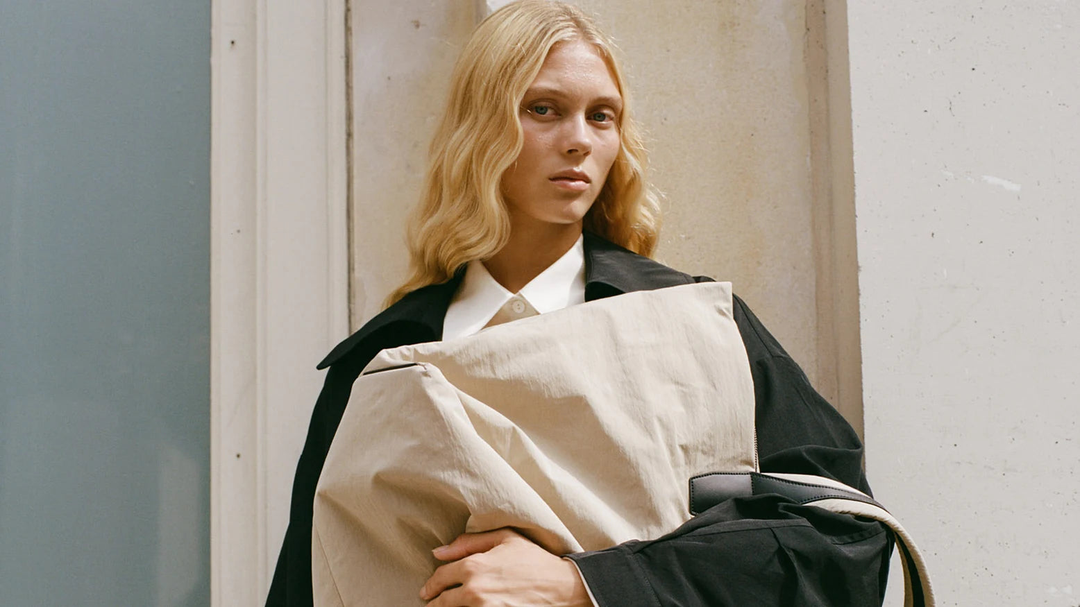 Woman in a black trench coat holding the Porter Essence Weekender Tote Bag in Beige Trench by Vee Collective in front of her upper body.