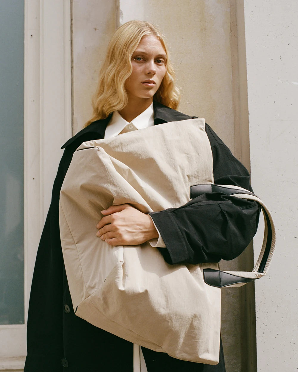 Woman in a black trench coat holding the Porter Essence Weekender Tote Bag in Beige Trench by Vee Collective in front of her upper body.