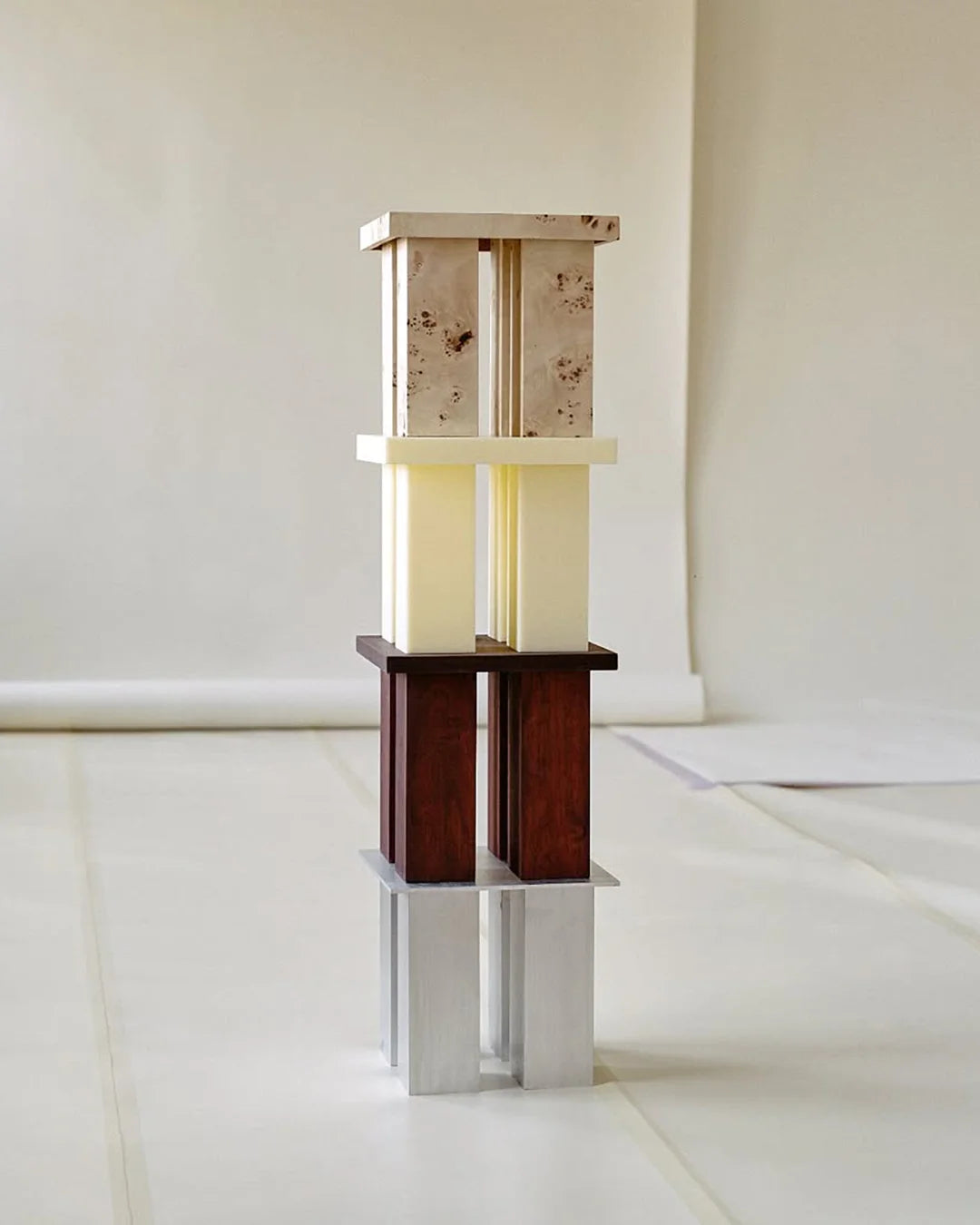 A tall, stacked sculptural object composed of rectangular blocks in varying materials and tones, including light stone, cream, dark wood, and brushed metal, arranged in a vertical column against a neutral studio backdrop.