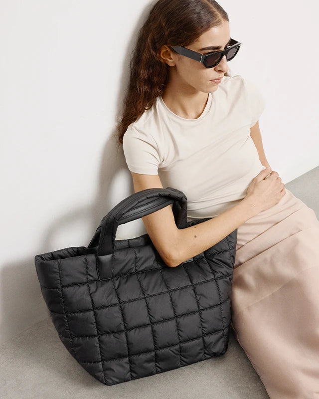 A woman wearing a beige outfit sits with the Porter Medium Tote Bag in black by Vee Collective positioned in next to her.