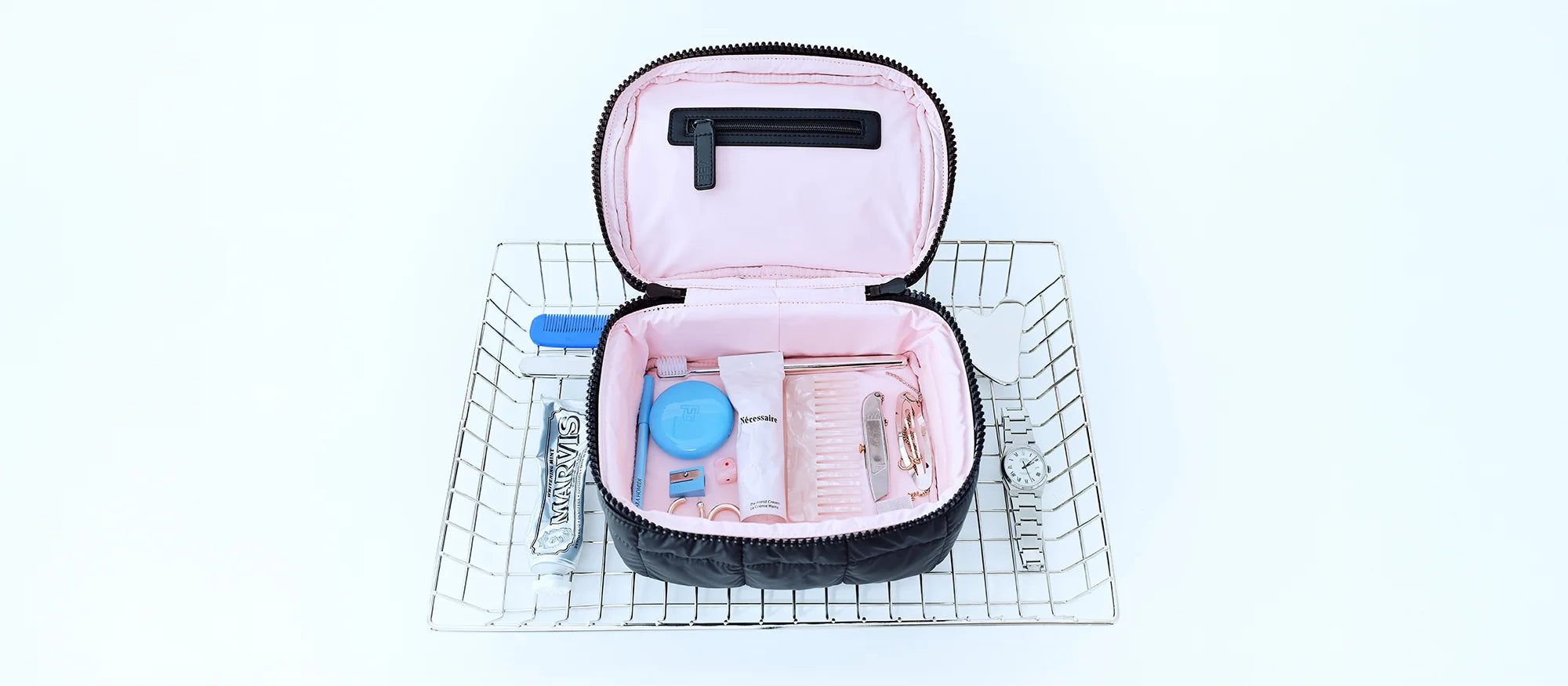 Top-down view of a basket with the Porter Vanity Case in matt black, opened to reveal its light pink interior, filled with cosmetics and jewelry, surrounded by additional cosmetic items.