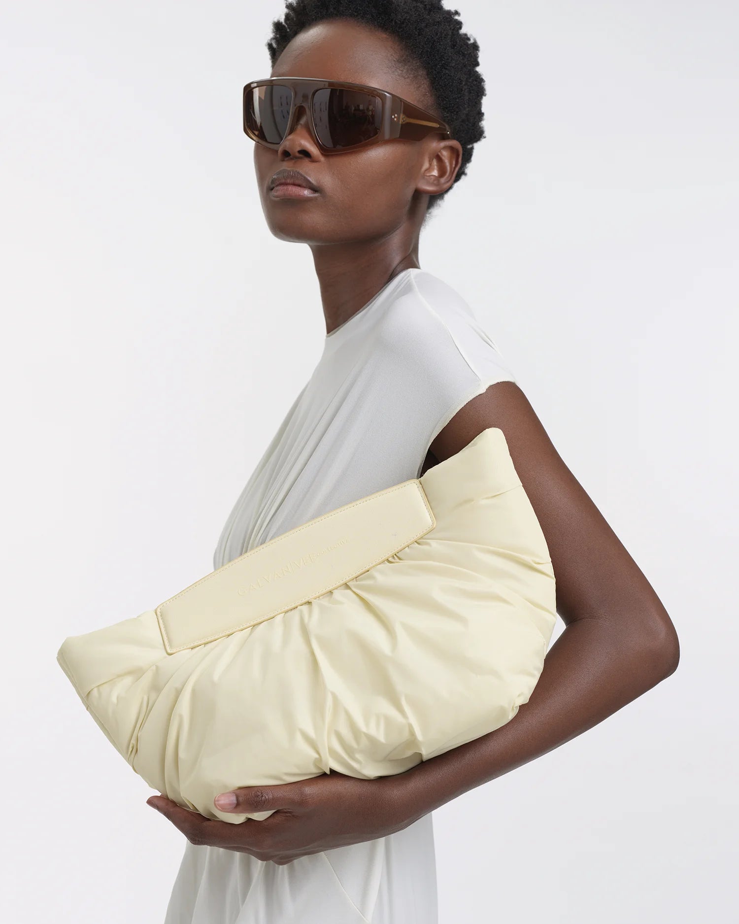 Woman standing in profile holding the Galvan x Vee Collective Caba Clutch in buttermilk in her arm, wearing a white dress.