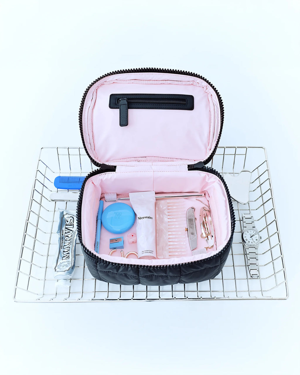 Bird’s-eye view of a basket showcasing the Porter Vanity Case in matt black by Vee Collective, open to display its light pink interior filled with cosmetics and jewelry, with various cosmetic items placed around it.