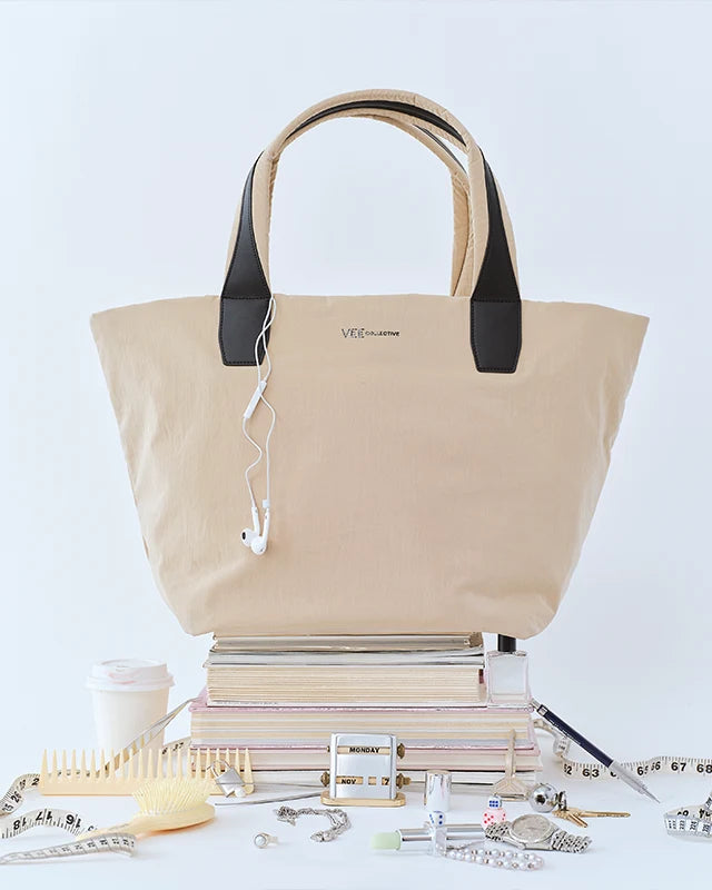 The Porter Essence Tote Bag Small in Beige Trench by Vee Collective displayed on a stack of books, with earphones hanging from one handle and various desk accessories arranged around it against a light background.