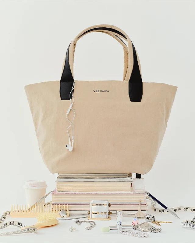 The Porter Essence Tote Bag Small in Beige Trench by Vee Collective displayed on a stack of books, with earphones hanging from one handle and various desk accessories arranged around it against a light background.