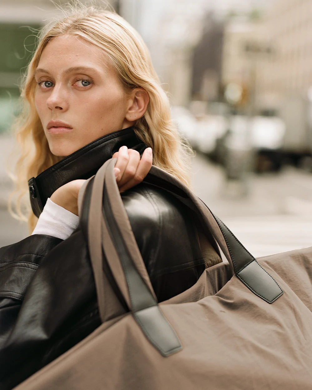 A woman with long blonde hair stands on a city street wearing a black leather jacket, carrying the Vee Collectives Porter Essence Weekender Tote Bag in Mink Trench over her shoulder.