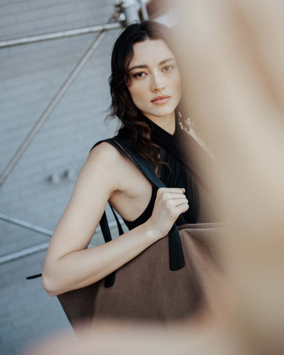 A woman wearing a black sleeveless top and black scarf, holding the Essence Ciy Tote Mink Trench with black handles, set against a gray brick wall background with a soft-focus foreground blur.
