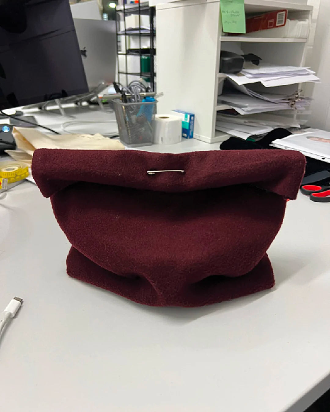 Prototype of the Galvan x Vee Collective Caba Clutch in mahogany, made of soft fabric and secured with a pin, placed on an office desk surrounded by papers and tools.