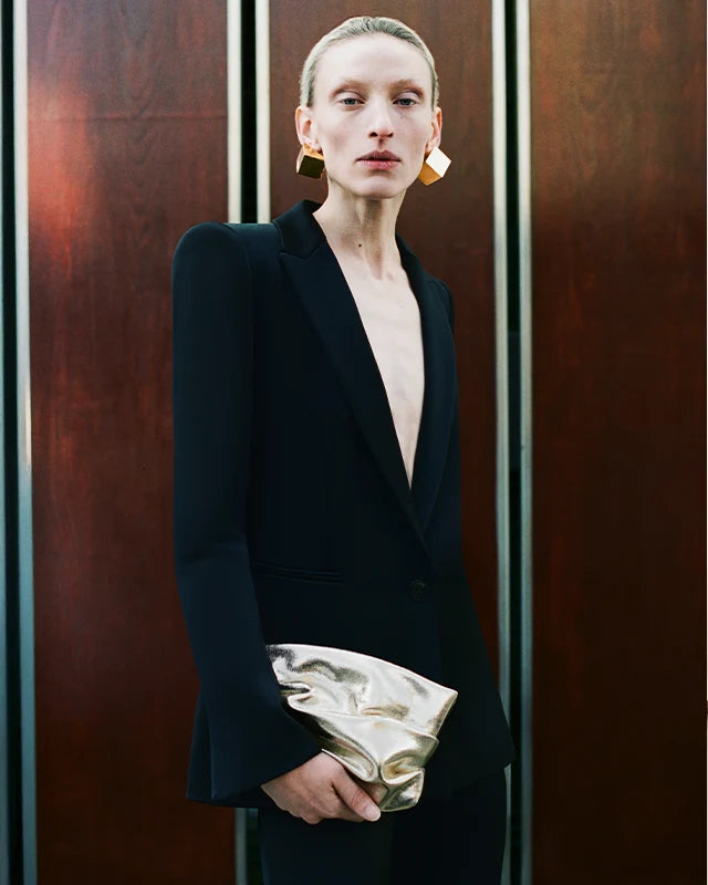 Woman wearing a structured black blazer and geometric gold earrings, holding the GALVAN x VEE Collective evening Clutch Bag in Gold Metallic in front of a wooden panel backdrop.