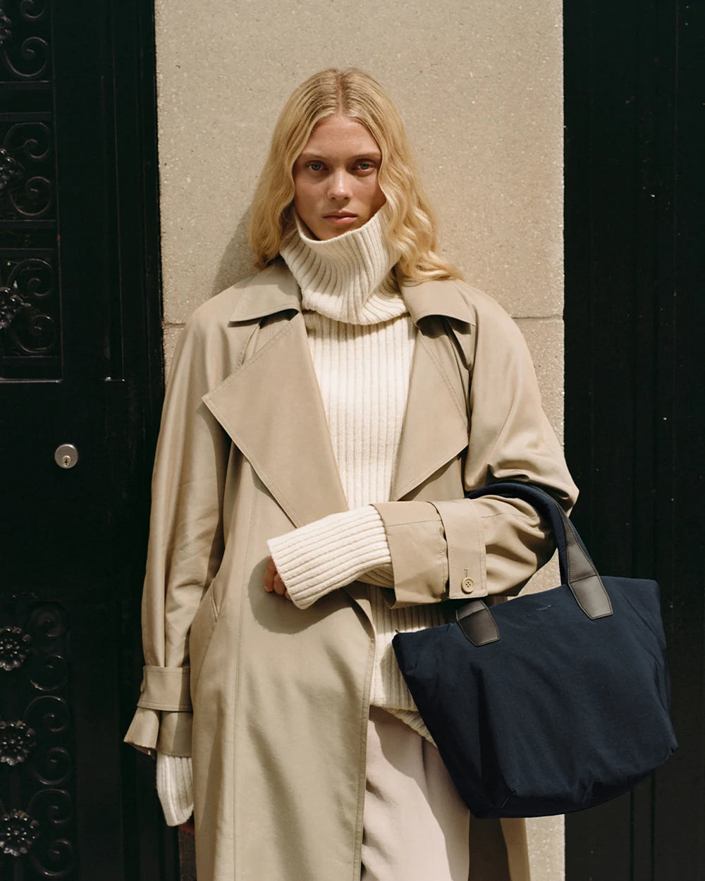 Woman in a beige trench coat holding the Porter Essence Small Tote Bag in Midnight Trench by Vee Collective against a plain studio background.