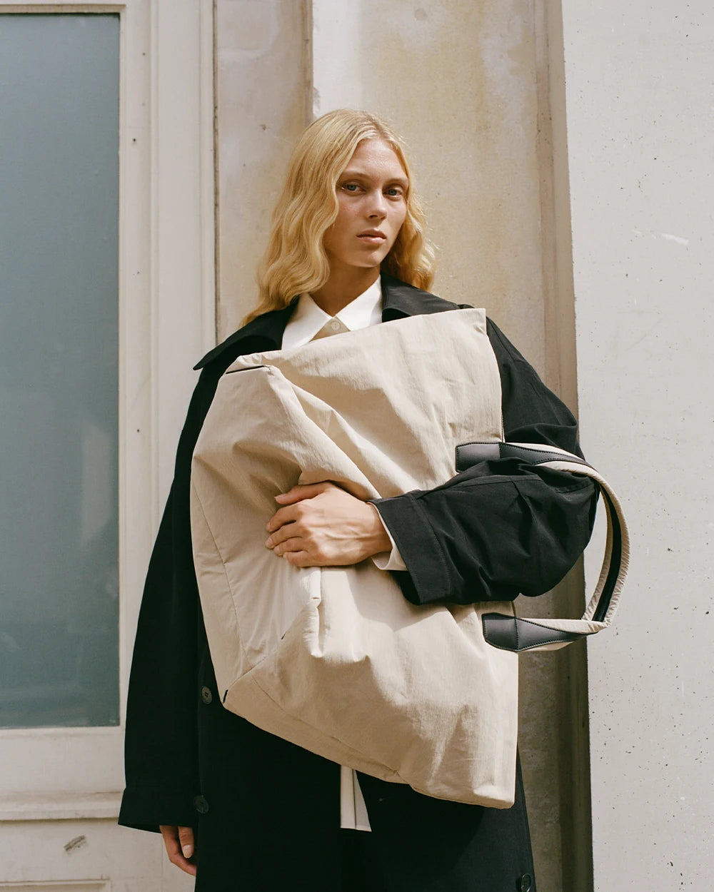 Woman in a black trench coat holding the Porter Essence Weekender Tote Bag in Beige Trench by Vee Collective in front of her upper body.