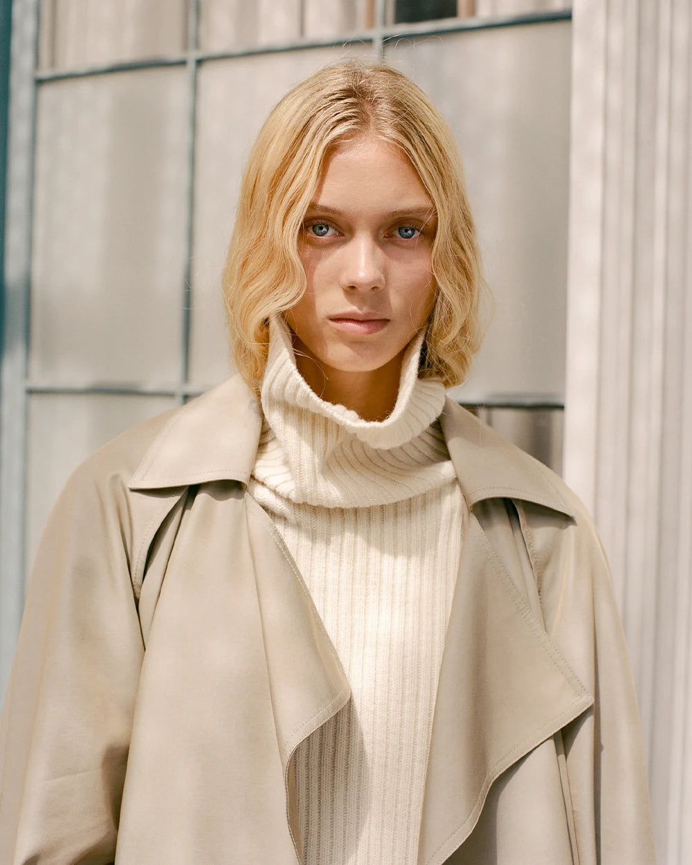 Woman in a beige trench coat standing against an urban background.
