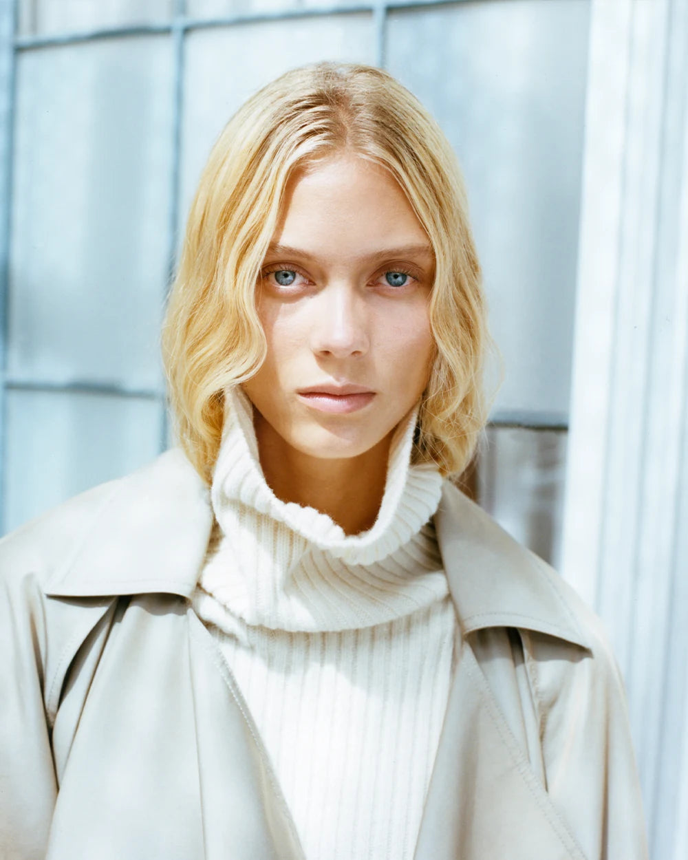 Woman with blond hair wearing a beige coat and white turtleneck sweater with a blurred background.
