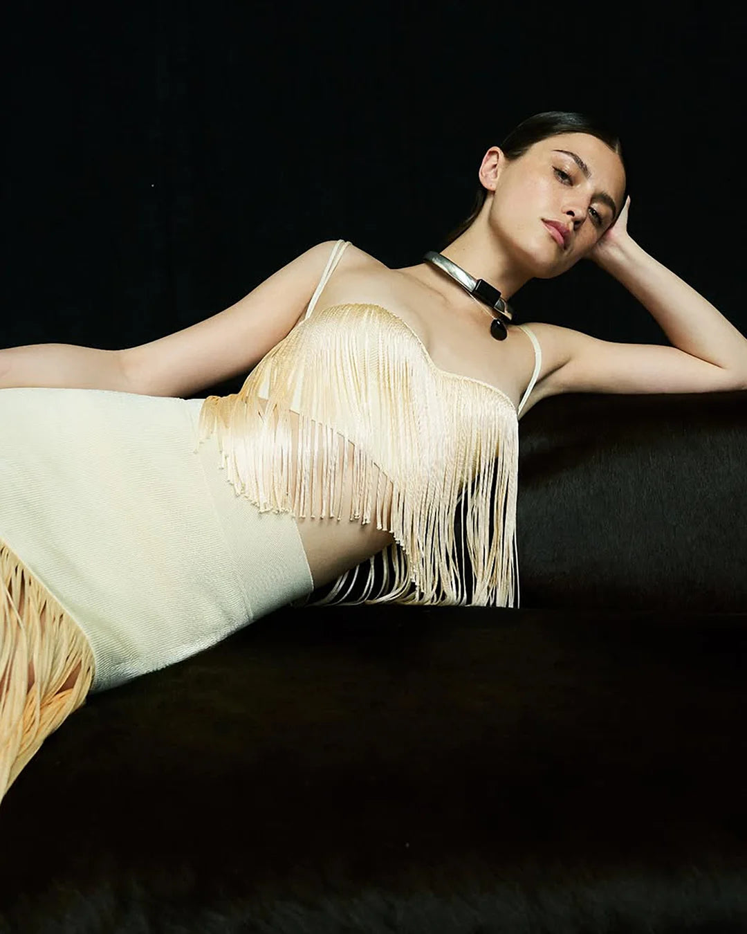 A woman wearing a cream-colored fringe top and matching skirt, with a sleek hairstyle and a bold metallic choker, set against a black background.