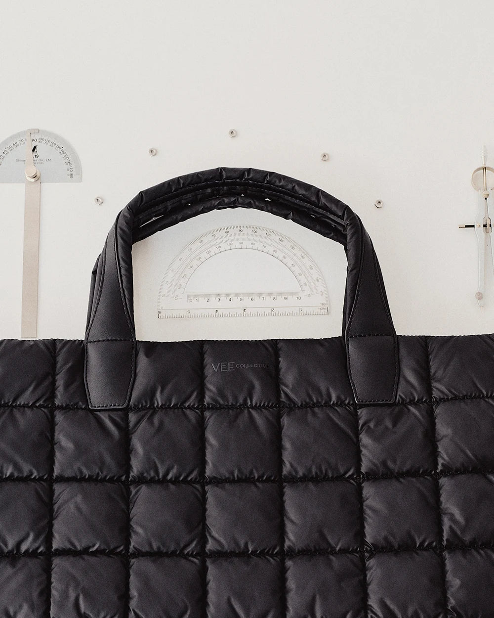 Black Porter Weekender Tote Bag by Vee Collective displayed against a white wall with protractors and measuring tools, highlighting its padded handles and modern geometric design