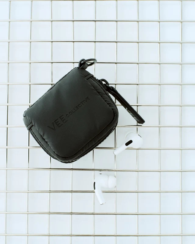 A Matt Black Vee Collective Porter Pods Case resting on a metal wire grid, shown with two white wireless earbuds placed nearby.