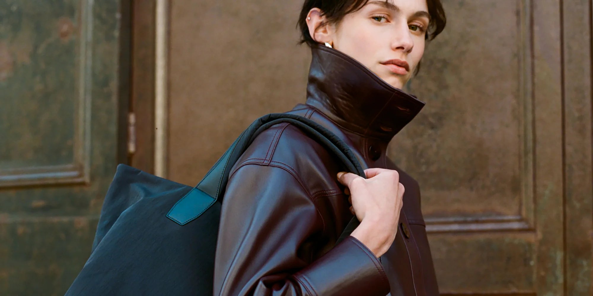 Woman wearing a brown jacket with a navy bag on her shoulder against a textured wall.