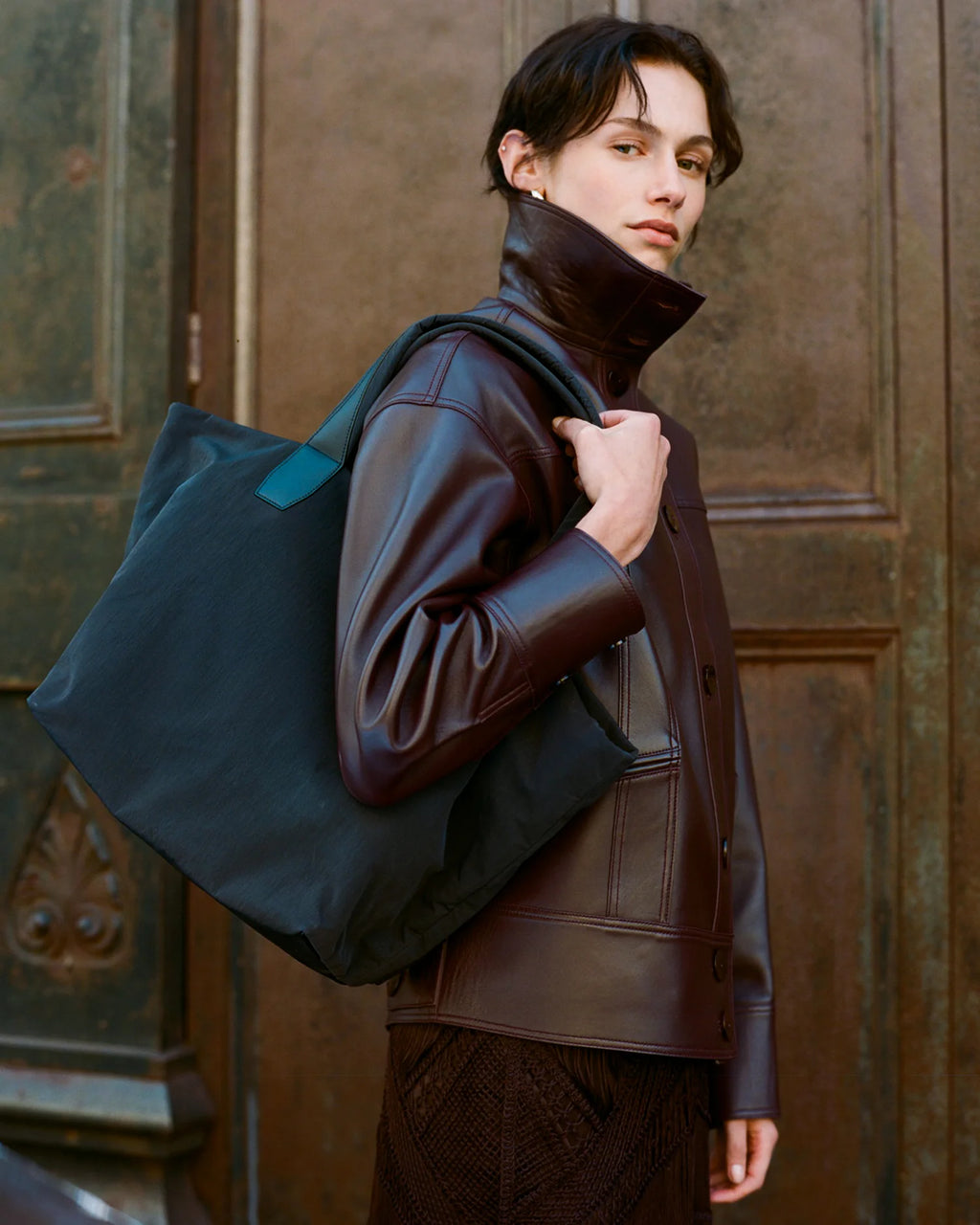 Woman wearing a brown leather jacket holding a navy  bag against a textured wall.