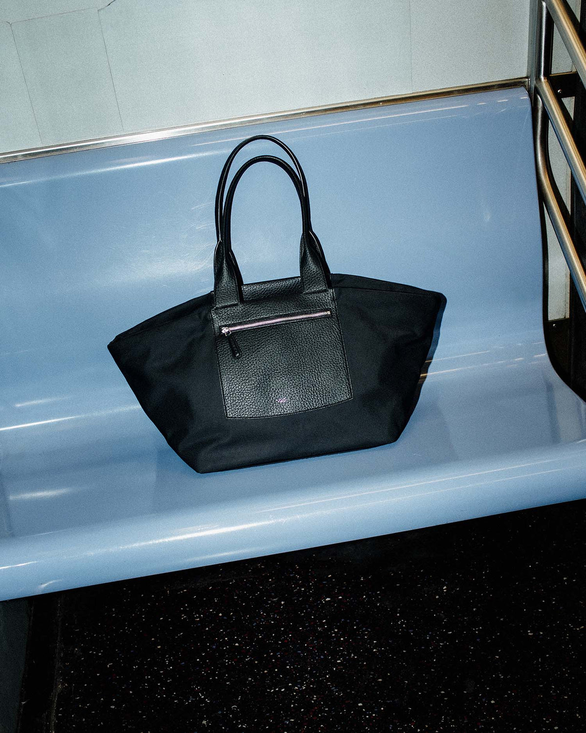 The black duo tote bag with dual top handles and a front zip pocket, resting on a light blue subway platform bench