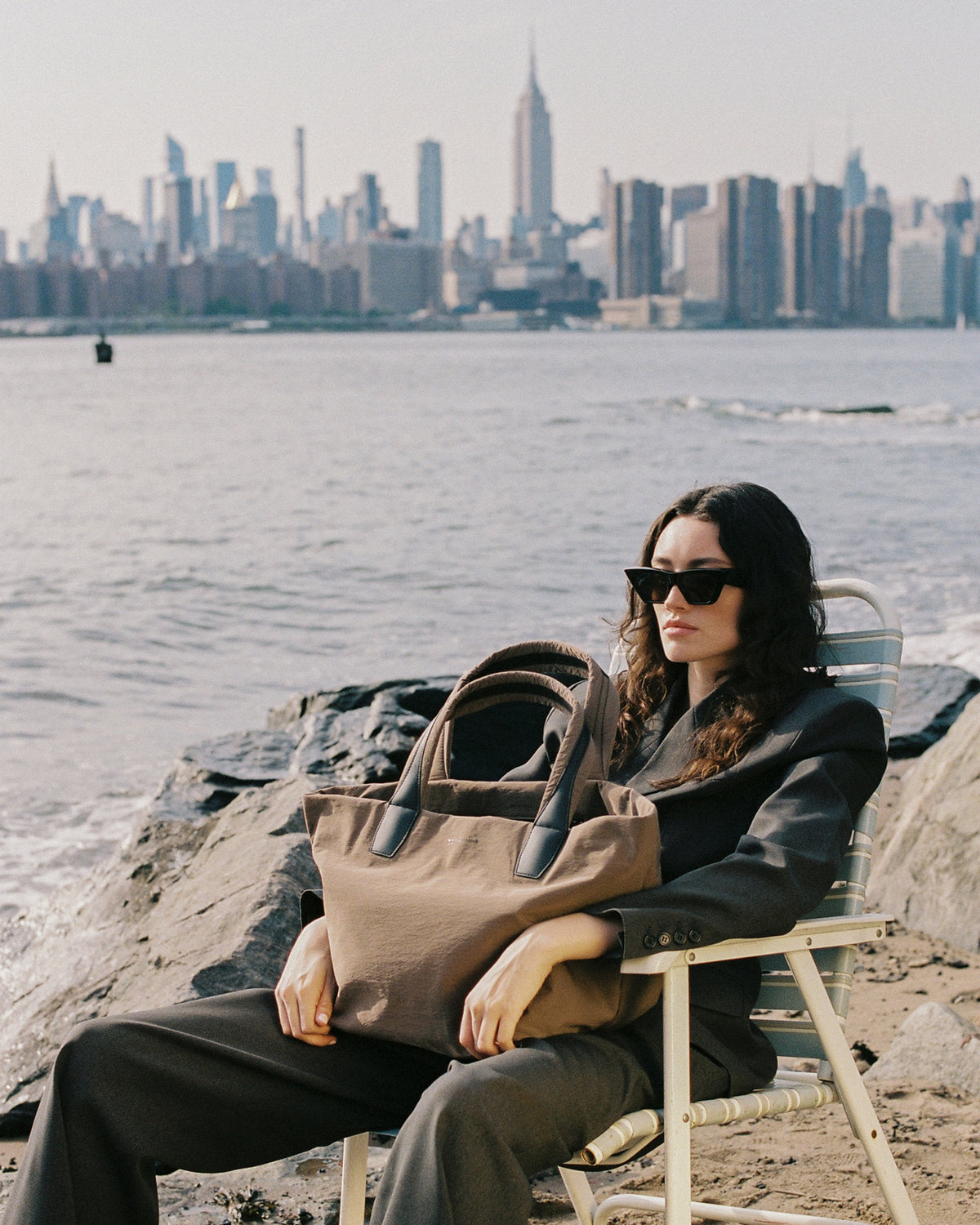 A photo of a model wearing black sunglasses and a dark blazer sits on a beach chair by the water, holding a large taupe canvas tote bag. The New York City skyline is visible in the background.