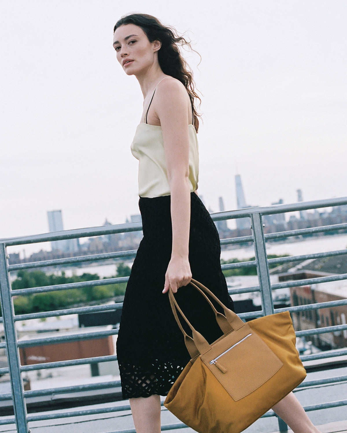 A Model with brown hair stands on an outdoor rooftop walkway carrying a large mustard yellow tote bag with tan leather handles and a front zip pocket. She wears a pale yellow camisole and a black skirt, with a city skyline and water in the background.