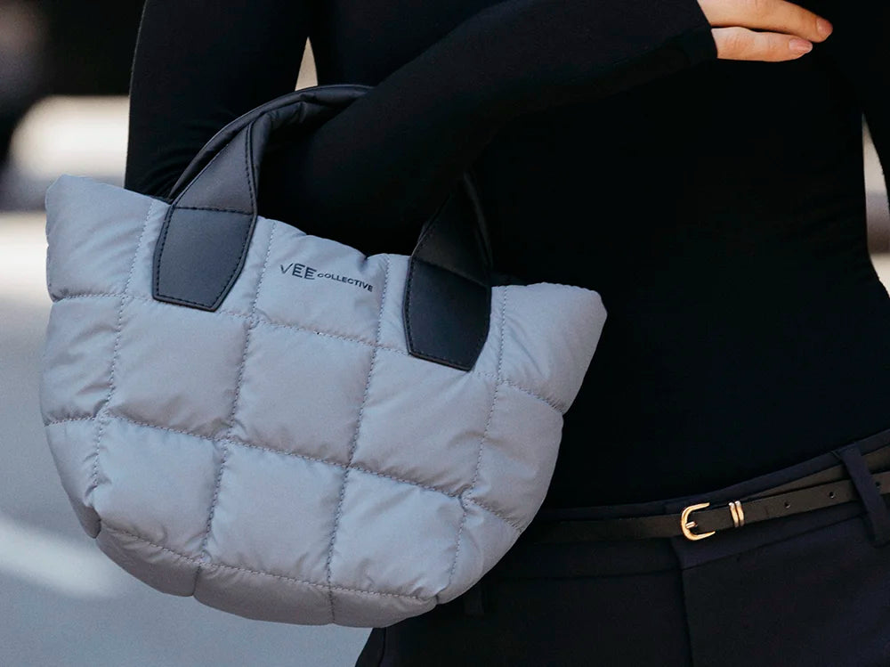 Gray quilted handbag with dark gray handles held by a person wearing a black outfit.