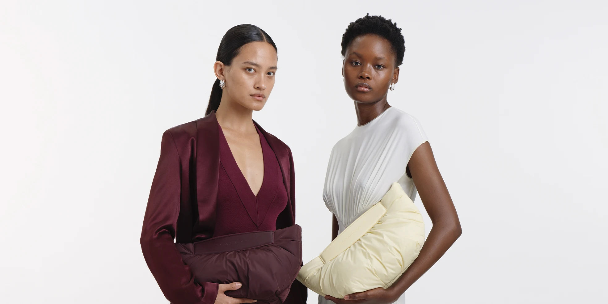 Fashion image of two models, one wearing a burgundy outfit with the matching Galvan x Vee Collective Caba Clutch in mahogany and the other in a white dress holding the Galvan x Vee Collective Caba Clutch in buttermilk, highlighting textures and colors.