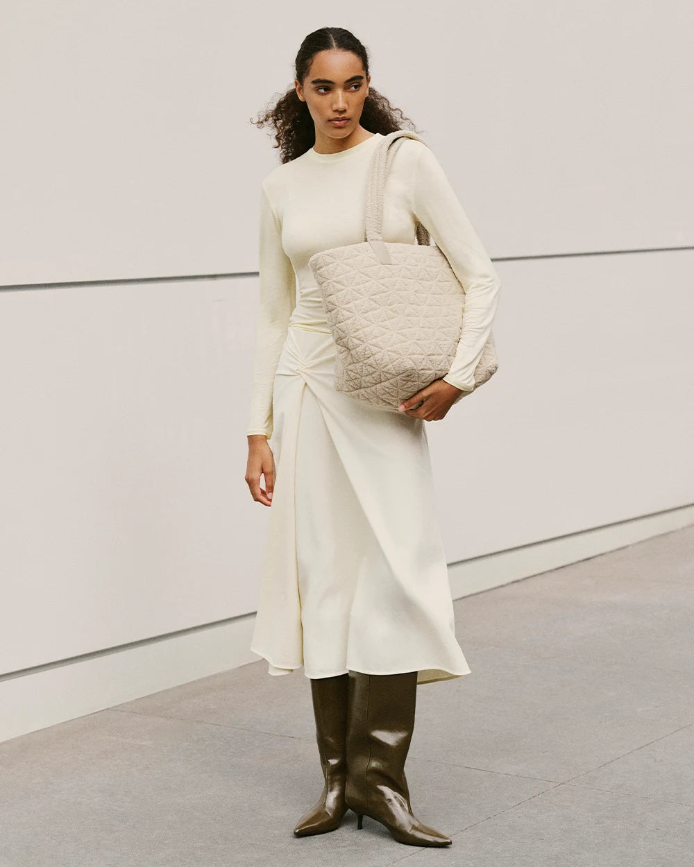 A woman stands with a Vee Tote Medium Cream Sherpa bag over her shoulder, wearing a beige dress outfit, in front of an urban background.
