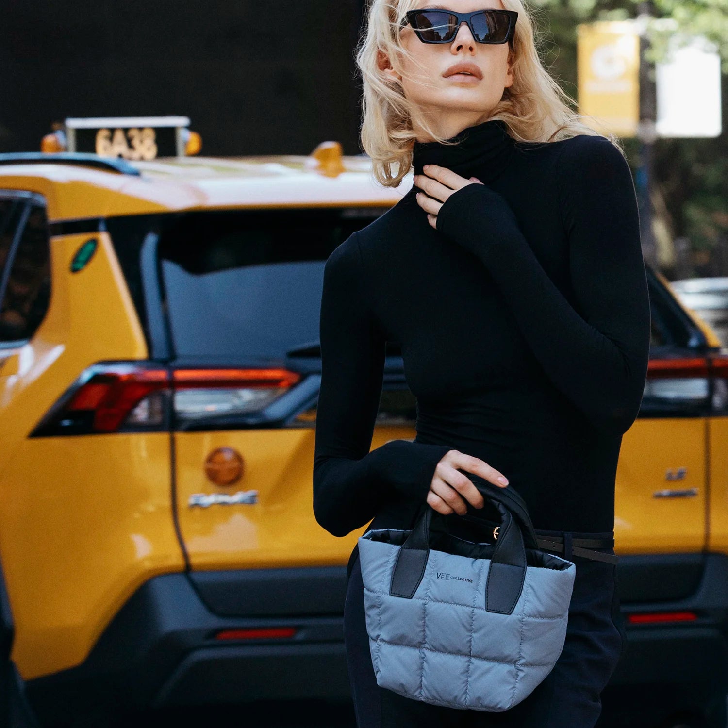 Woman in black outfit holding a gray quilted handbag with a yellow taxi in the background