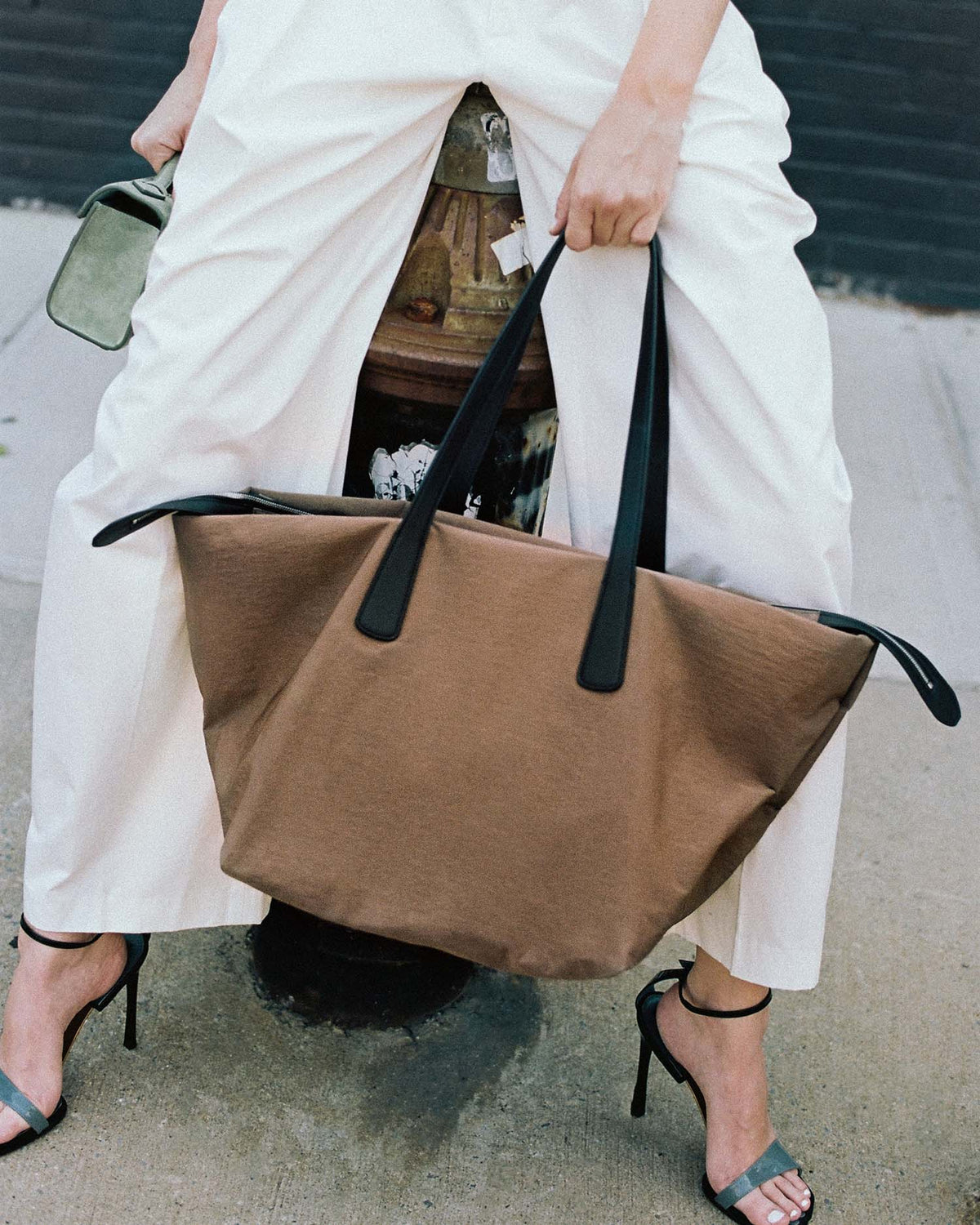A person wearing white pants and black high heels carries a Essence City Tote Large Mink Trench bag with black handles on a city sidewalk.