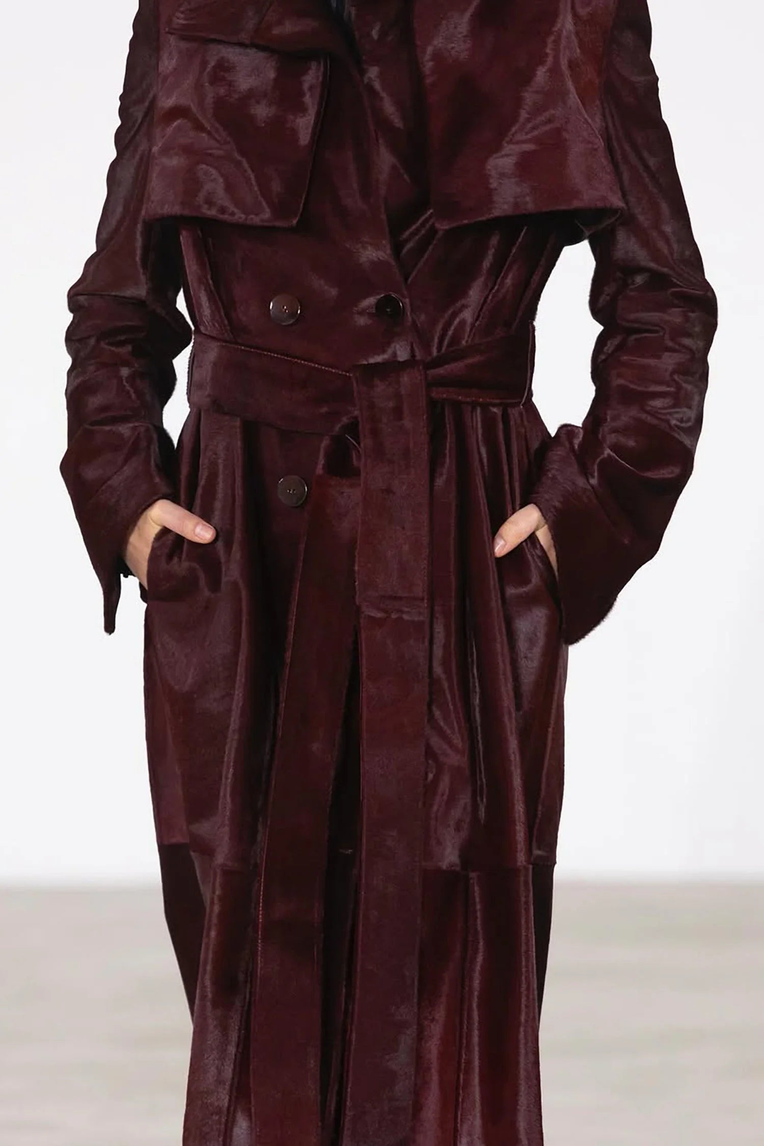 Close-up of a woman wearing a deep mahogany long coat with a soft, glossy texture. The coat features a wide lapel, double-breasted buttons, and a tied waist belt, with her hands placed in the side pockets.