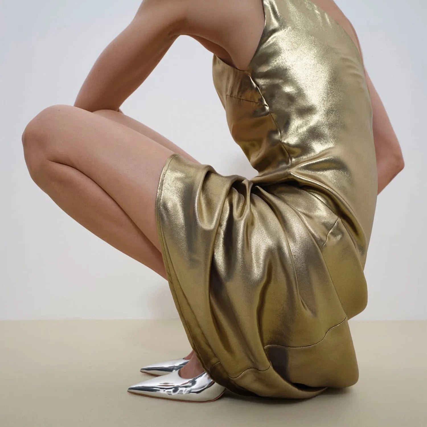 A person crouches down wearing a metallic gold slip dress and pointed silver heels. The image focuses on the draped texture of the dress and the reflective finish of the shoes, with a minimal neutral background.