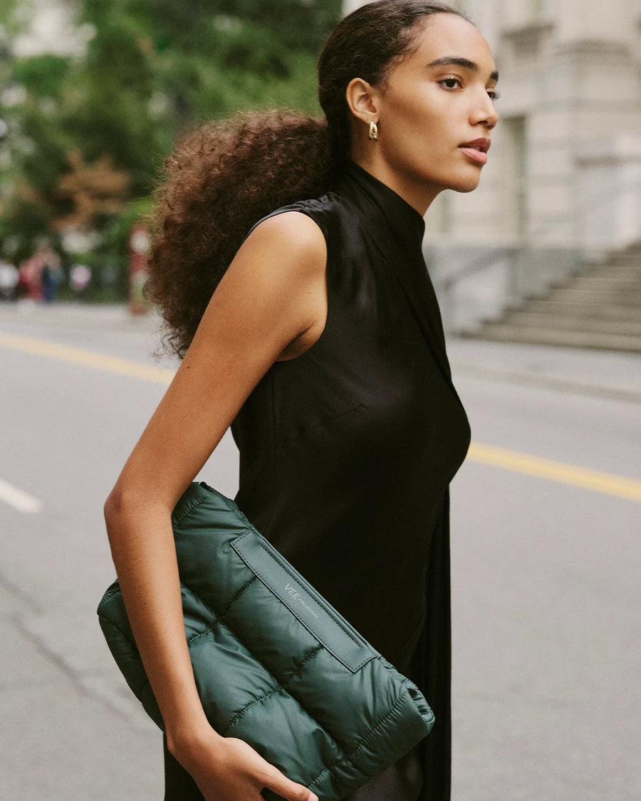 Close-up of a woman holding the Vee Collective Porter Basque Clutch in Bottle Green, styled with a neutral outfit in an urban street setting.