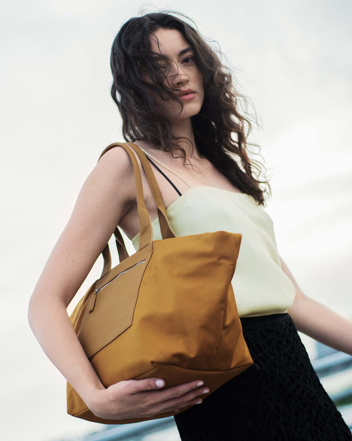 A model carrying a large mustard yellow canvas tote bag with brown leather handles and a brown leather front pocket, styled with a pale yellow top and a black skirt against a bright, overcast sky.