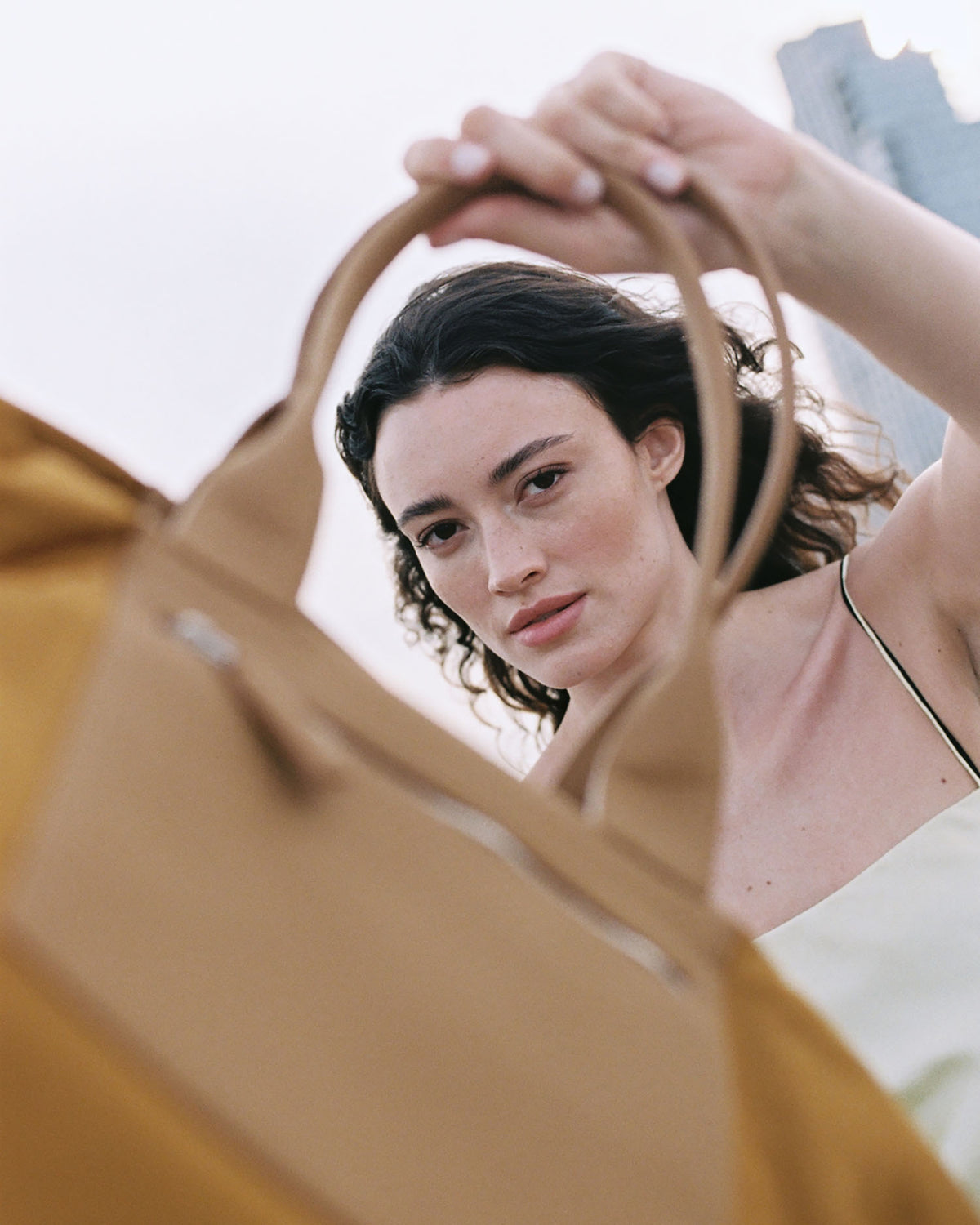 A close-up outdoor photo featuring a model holding a Vee Ta Tracy Anderson tote bag, with the bag’s handles framing part of her face and upper body.