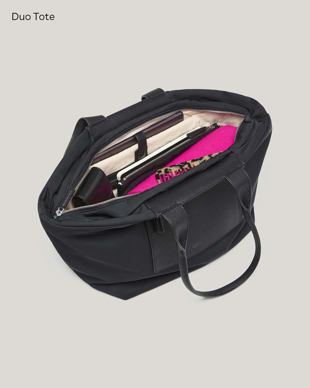 An open black Duo Tote bag in medium shown from above on a white background, revealing a cream interior organizer with dark accessories and a bright pink item inside.