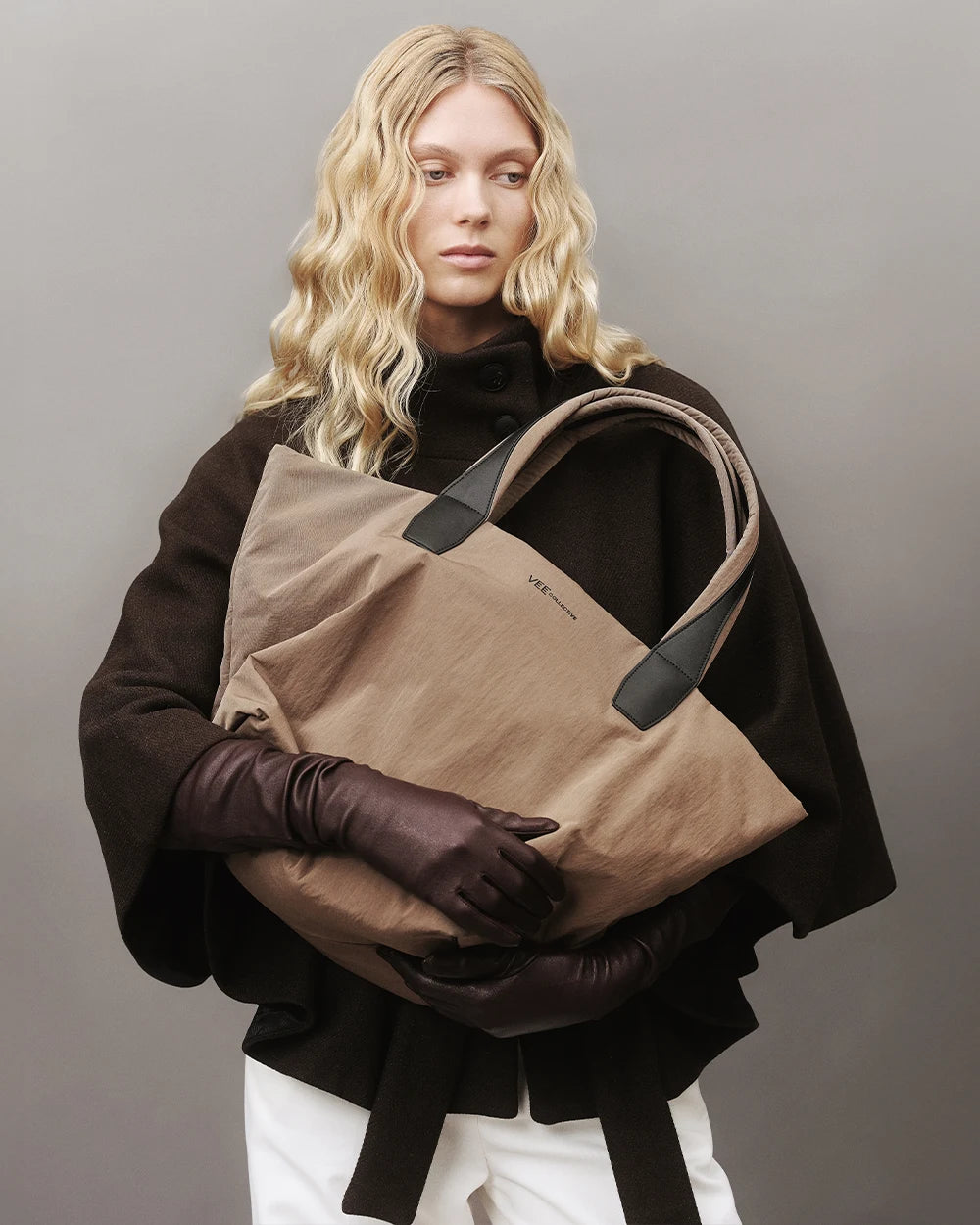 Model with blonde hair wearing a dark brown coat and matching leather gloves, holding the Vee Collective Essence Tote Medium bag in Mink Trench against a gray background.