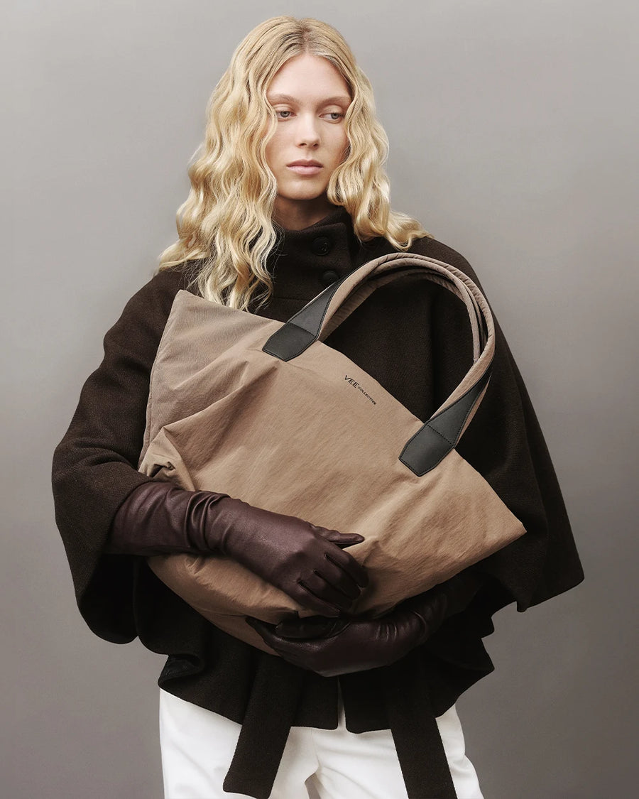 Model with blonde hair wearing a dark brown coat and matching leather gloves, holding the Vee Collective Essence Tote Medium bag in Mink Trench against a gray background.