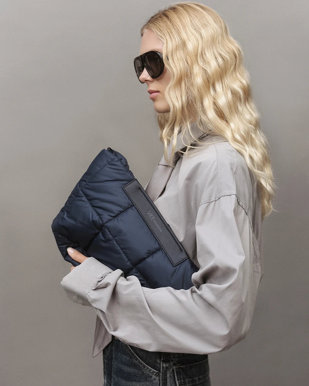 Model with blonde hair wearing a gray shirt, jeans, and sunglasses, holding the Vee Collective Porter Basque Clutch in Dark Navy against a gray background.