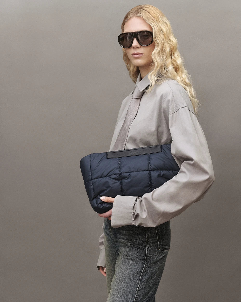 Model with blonde hair wearing a gray shirt, jeans, and sunglasses, holding the Vee Collective Porter Basque Clutch in Dark Navy against a gray background.