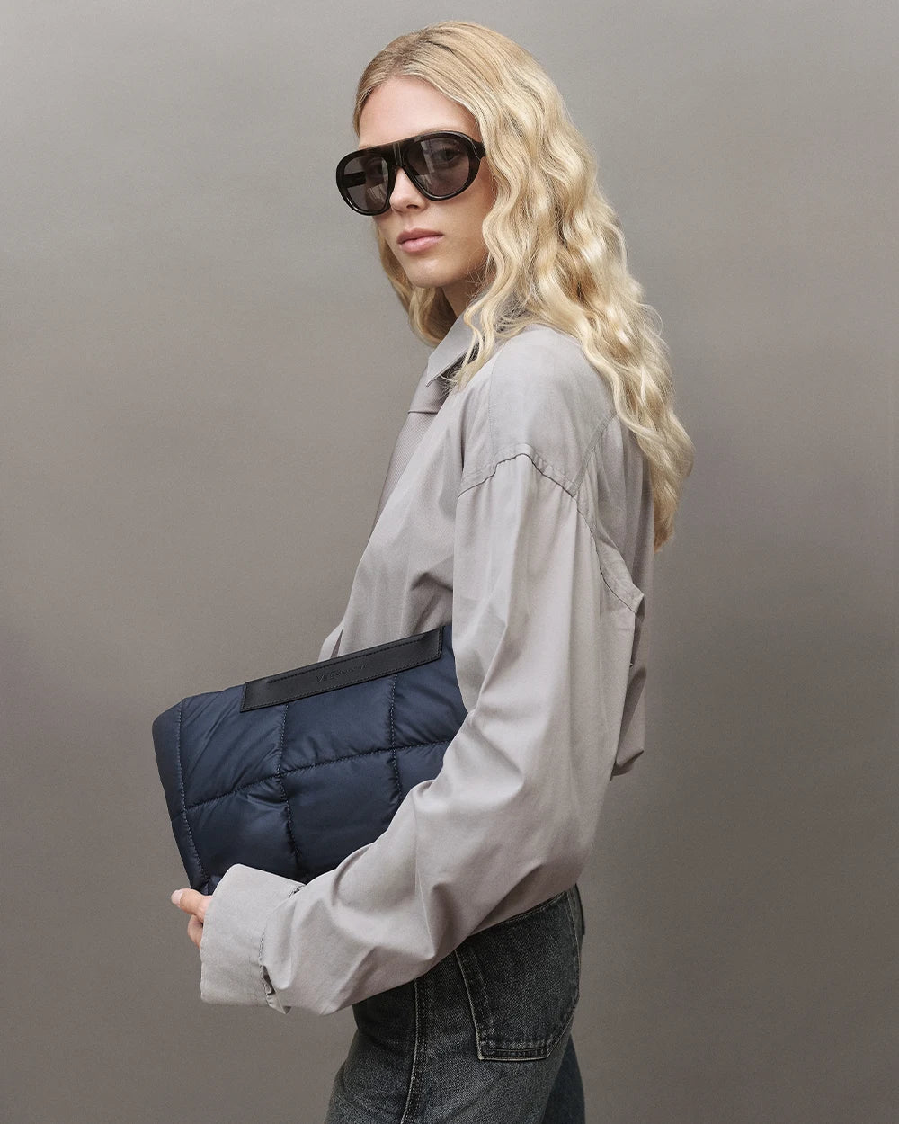 Model with blonde hair wearing a gray shirt, jeans, and sunglasses, holding the Vee Collective Porter Basque Clutch in Dark Navy against a gray background.