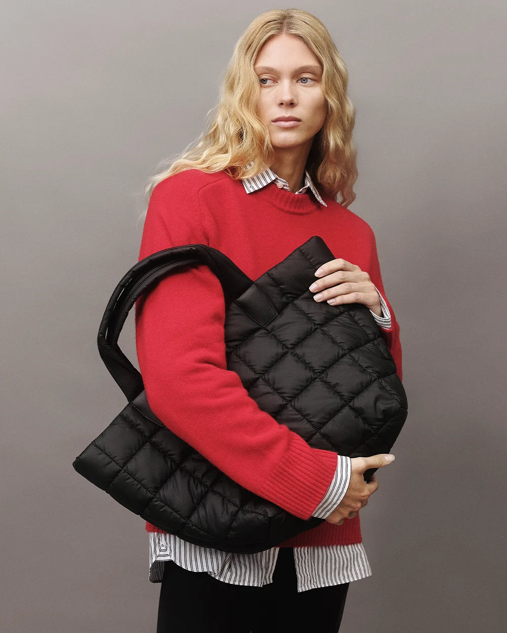 Model with blonde hair wearing a striped shirt, red jumper, and black pants, holding the Vee Collective Porter Weekender bag in Matt Black against a gray background.