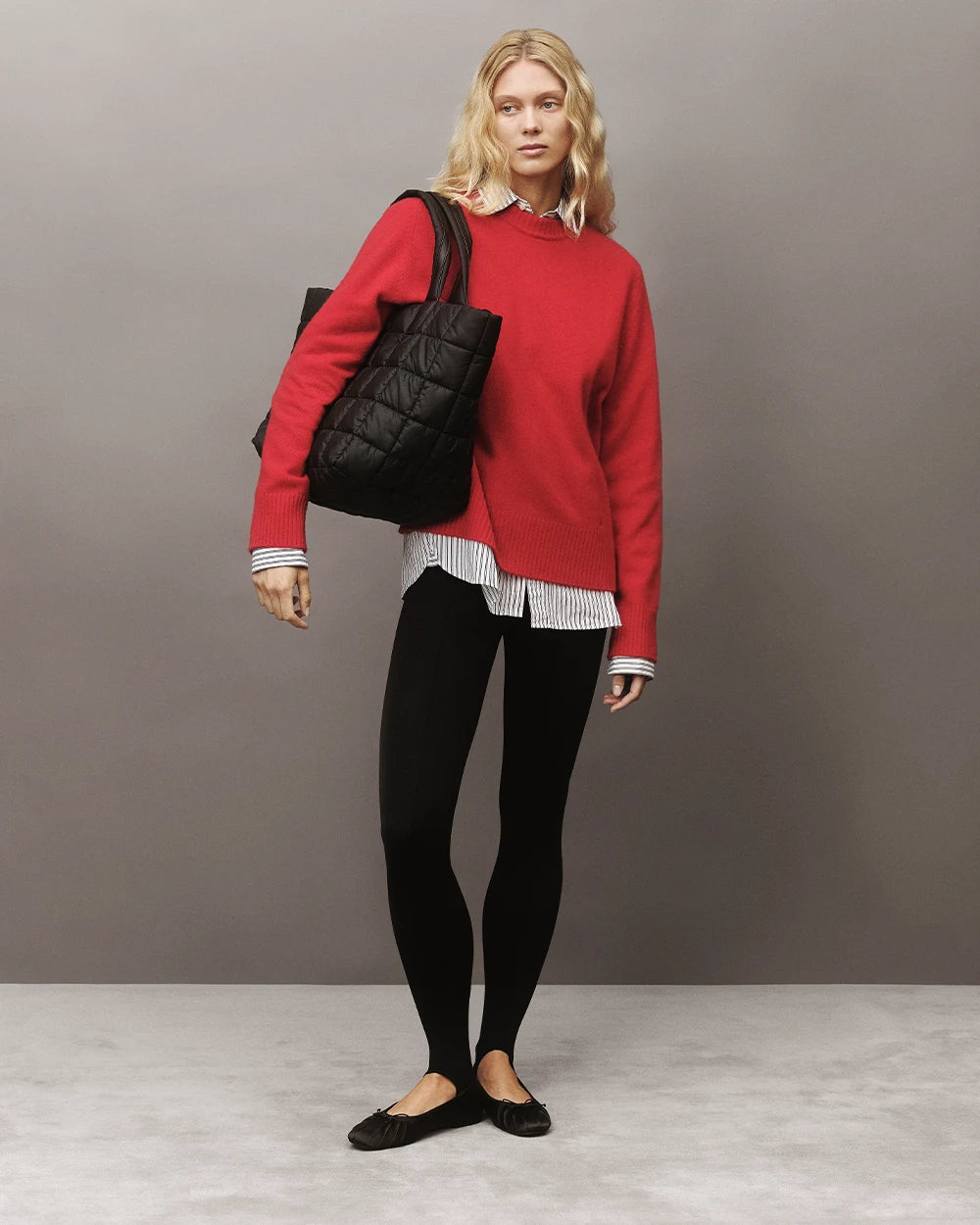Model with blonde hair wearing a striped shirt, red jumper, black pants and matching flat shoes, holding the Vee Collective Porter Weekender bag in Matt Black against a gray background.