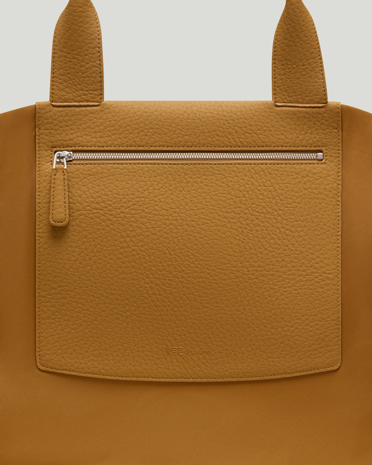 Close-up of the Duo Tote large honey mustard tote bag showing a front zip pocket with a silver zipper and a small hanging pull tab, set against a white  background.