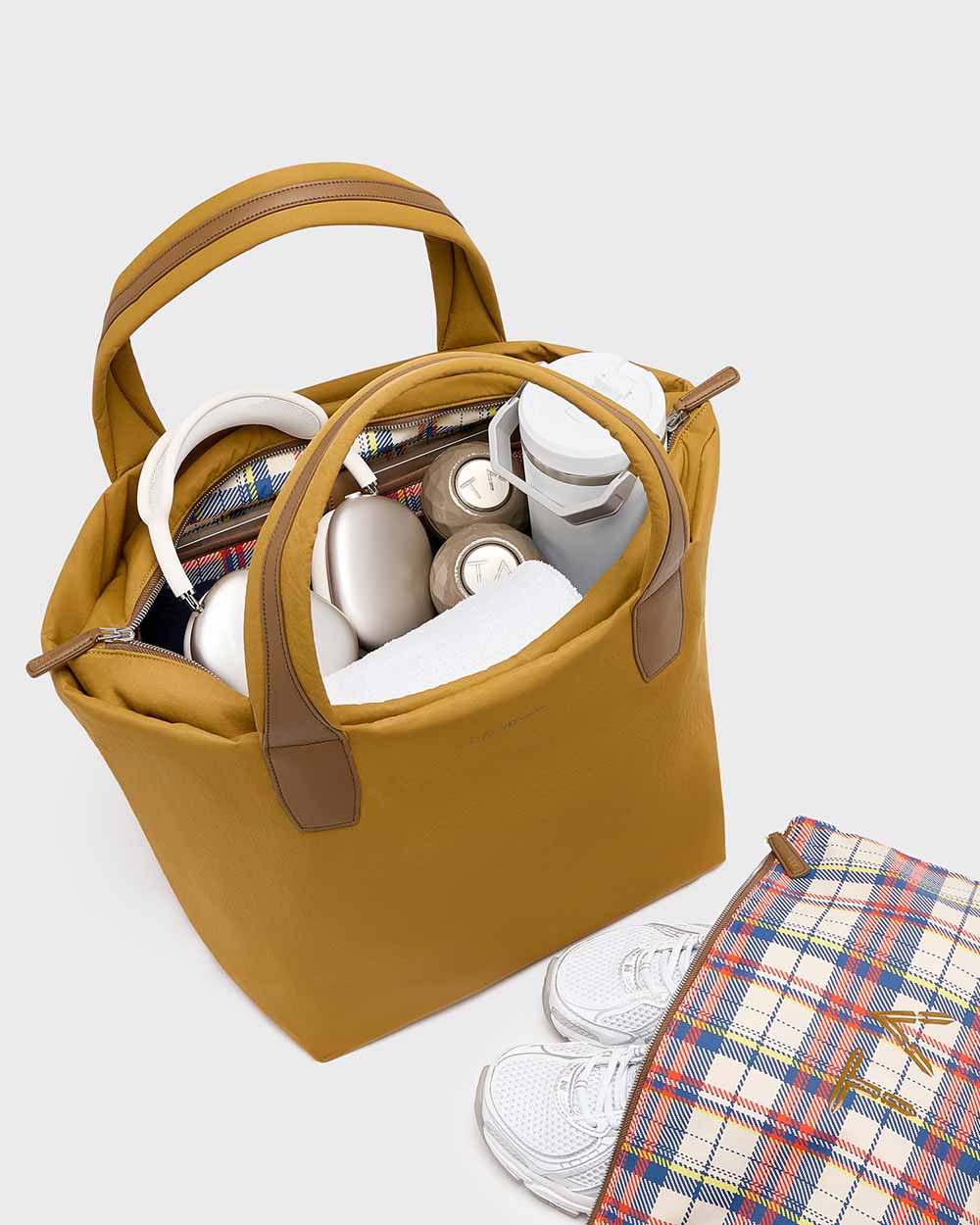 The Essence Tote Medium Tracy Anderson  bag with brown handles, shown open and filled with travel items including a water bottle, cup, and small accessories. A plaid zip pouch and white sneakers are placed beside it on a white  background.