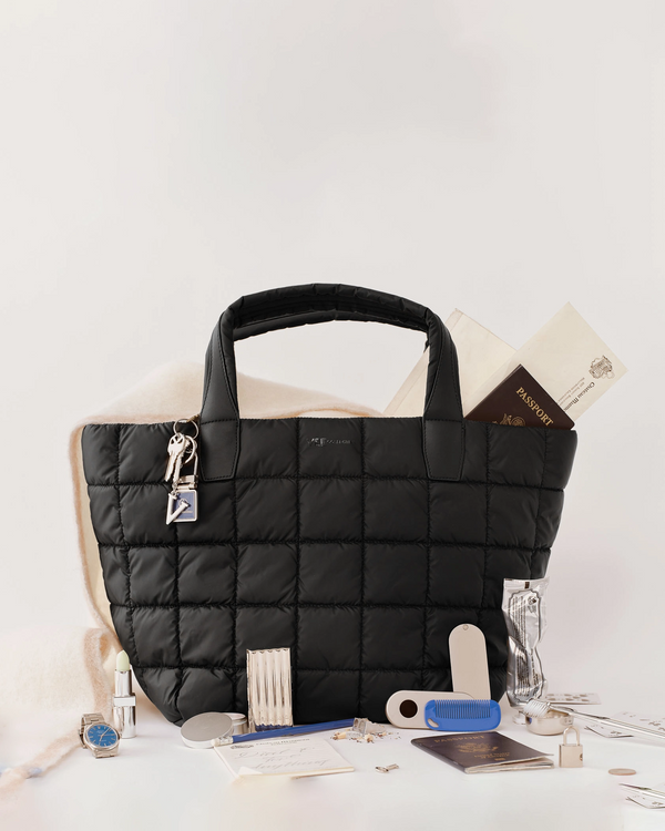 A black Porter Tote bag with silver-tone hardware and a hanging lock, styled on a white surface with travel accessories including a passport, perfume bottle, wallet, and keys.