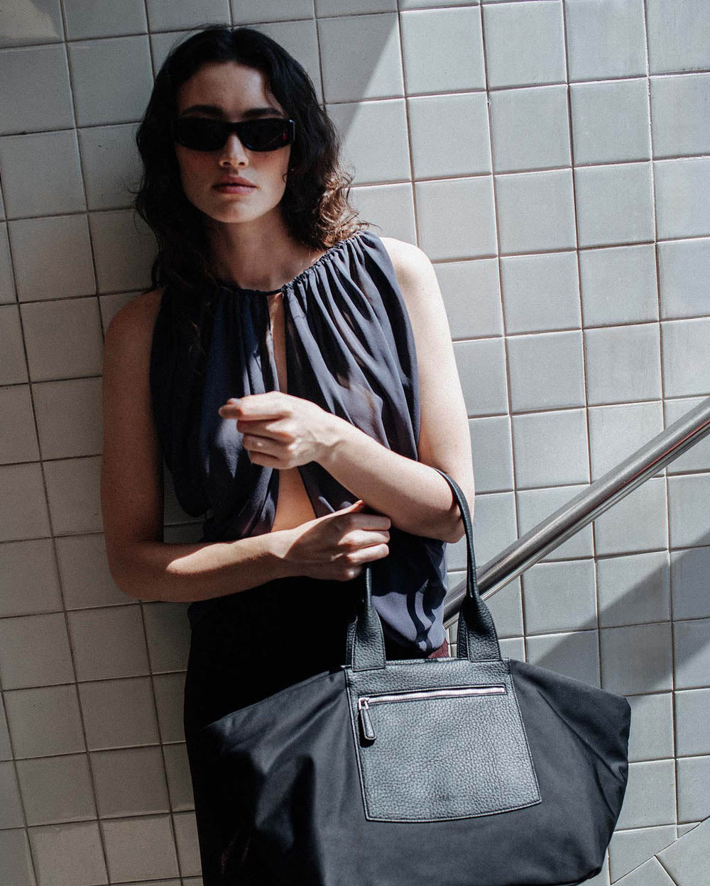 A photo featuring a model wearing black sunglasses and a sheer black sleeveless top stands against a tiled wall, carrying a large black Duo Tote Bag with a leather front pocket.